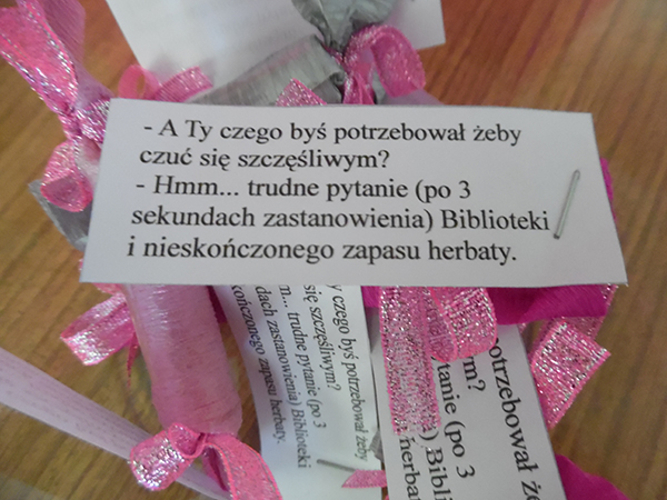 Zdjęcie kartki z cytatem na tle różowych wstążeczek. ,,-A Ty czego byś potrzebował żeby czuć się szczęśliwym? -Hmm... trudne pytanie (po 3 sekundach zastanowienia) Biblioteki i nieskończonego zapasu herbaty."