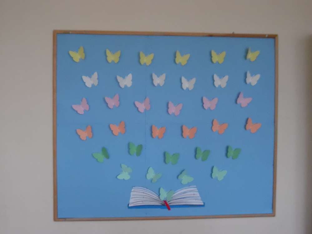 Tablica okolicznościowa wykonana w bibliotece w celu promowania czytelnictwa wśród uczniów. Na błękitnym tle, na samym dole, w centralnym miejscu znajduje się otwarta książka wykonana z papieru. Z książki „wylatują” papierowe motyle w pastelowych kolorach.