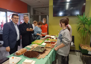 Burmistrz Rzgowa, ks. proboszcz- kanonik oraz członkowie PZ CARITAS przy stole ze słodkimi wypiekami.