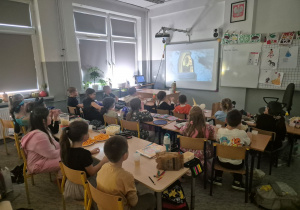 Na zdjęciu uczniowie klasy 1a siedzą w ławkach, na których znajdują się miseczki z przekąskami, takimi jak popcorn i chrupki. Dzieci z zainteresowaniem oglądają film wyświetlany na tablicy interaktywnej.