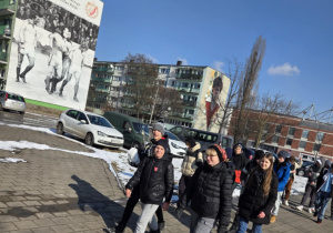 Uczestnicy idą wzdłuż bloków przy al. Piłsudskiego, na fasadach bloków widać murale nawiązujące do historii kluby, m.in. Włodzimierz Smolarek i Zbigniew Boniek