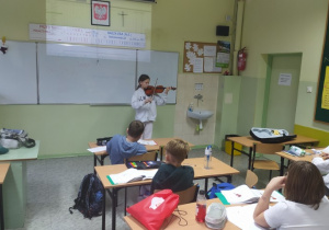 Uczennica gra na skrzypcach etiudę, uczniowie słuchają z zaciekawieniem