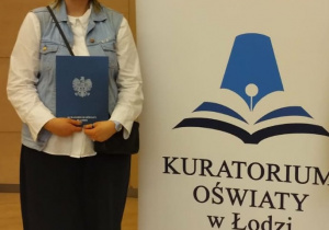 Alicja Pietraszczyk z dyplomem na tle baneru Kuratorium Oświaty w Łodzi