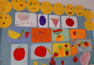 Galeria prac dzieci zaprezentowana na tablicy korkowej. Na górze żółte buźki przedstawiające emocje (stały element dekoracji). Pozostała przestrzeń to wycinanki z kolorowego papieru: owoce i warzywa.