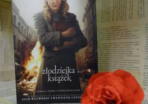 Zdjęcie przedstawia książkę ustawioną na regale wystawowym w bibliotece. Jest to książka, pt. „Złodziejka książek” Markusa Zusaka. Obok książki znajduje się kwiat, wykonany z czerwonej bibuły.