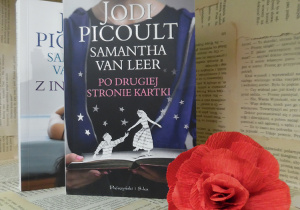 Zdjęcie przedstawia dwie książki amerykańskiej powieściopisarki Jodi Picoult, pt. „Z innej bajki” i „Po drugiej stronie kartki”. Książki zostały ustawione na regale wystawowym w bibliotece. Obok nich znajduje się kwiat, wykonany z czerwonej bibuły.