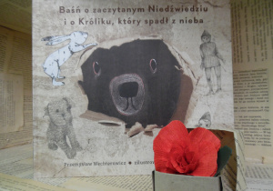 Zdjęcie przedstawia książkę pt. „Baśń o zaczytanym Niedźwiedziu i o Króliku, który spadł z nieba” Przemysława Wechterowicza, ustawioną na regale wystawowym w bibliotece. Obok książki znajduje się małe, papierowe pudełko z kwiatem wykonanym z czerwonej bibuły.