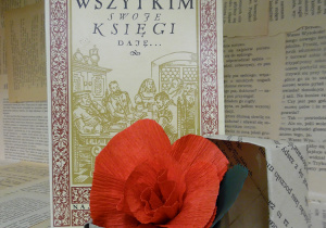 Zdjęcie przedstawia książkę Miry Jaworczakowej, pt. „Wszytkim swoje księgi daję...”, ustawioną na regale wystawowym w bibliotece. Obok książki znajduje się małe, papierowe pudełko z kwiatem, wykonanym z czerwonej bibuły.