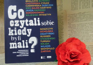 Zdjęcie przedstawia książkę Ewy Świerżewskiej i Jarosława Mikołajewskiego, pt. „Co czytali sobie kiedy byli mali?”, ustawioną na regale wystawowym w bibliotece. Obok książki znajduje się kwiat, wykonany z czerwonej bibuły.