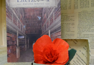 Zdjęcie przedstawia książkę Pauliny Mikuły, pt. „Literatoura”, ustawioną na regale wystawowym w bibliotece. Obok książki znajduje się małe, papierowe pudełko z kwiatem, wykonanym z czerwonej bibuły.