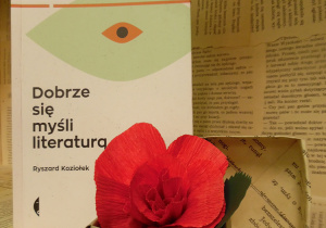 Zdjęcie przedstawia książkę napisaną przez Ryszarda Koziołka, pt. „Dobrze się myśli literaturą”, ustawioną na regale wystawowym w bibliotece. Obok książki znajduje się małe, papierowe pudełko z kwiatem, wykonanym z czerwonej bibuły.