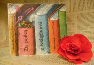 Zdjęcie przedstawia książkę pt. „Za półką z książkami” Jolanty Hartwig-Sosnowskiej, ustawioną na regale wystawowym w bibliotece. Obok książki znajduje się kwiat, wykonany z czerwonej bibuły.