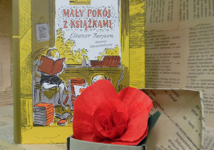 Zdjęcie przedstawia książkę ustawioną na regale wystawowym w bibliotece. Jest to pozycja napisana przez Eleanor Farjeon, brytyjską pisarkę i poetkę. Obok książki pt. „Mały pokój z książkami” znajduje się małe, papierowe pudełko z kwiatem, wykonanym z czerwonej bibuły.