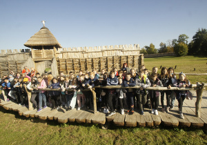 Duża grupa uczniów stojąca na drewnianej drodze przed bramą prehistorycznej osady w Biskupinie.