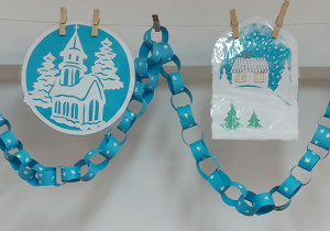 Zimowe witraże wycięte z białego papieru, podklejone gładka bibułą. Łańcuch świąteczny wykonany przez dzieci z niebieskiego kartonu i białego korektora.