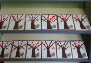 Zdjęcie przedstawia książki dla uczniów klas I, ustawione na regale bibliotecznym. Każdy egzemplarz ozdobiony jest drzewkiem w czerwonymi serduszkami, oznaczeniem klasy i numerem książki.