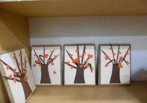 Zdjęcie wykonane w ramach zabawy bibliotecznej Randka w ciemno z książką - VIII EDYCJA. Na półce regału znajdują się cztery opakowane książki. Wierzch każdej z nich zdobi drzewo z czerwonymi serduszkami.