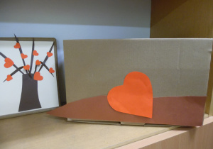 Zdjęcie wykonane w ramach zabawy bibliotecznej Randka w ciemno z książką - VIII EDYCJA. Na półce regału znajduje się książka, ozdobiona drzewkiem z czerwonymi serduszkami i brązowe pudełko na karty książek.