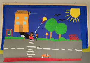 Dekoracja ścienna przedstawiająca ruch uliczny: budynek szkoły, pojazdy, sygnalizacja świetlna, dziewczynka, drzewa, znaki drogowe. Praca wykonana w gr. III