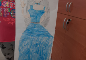 Dziewczynka trzymająca swoją pracę z rysunkiem księżniczki w niebieskiej sukni. Praca wykonana w gr. I pod opieką Pani Pauli Filipczak.
