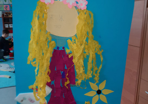 Duży plakat przedstawiający Panią Wiosnę. Kobieta ma fioletową suknię, jasne włosy i kolorowy wianek z kwiatów. Praca wykonana w gr. I pod opieką Pani P. Filipczak.